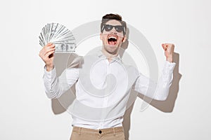 Emotional brunette guy in shirt and sunglasses shouting with clenched fist while holding money prize in cash, isolated over white