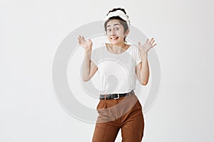 Emotional clueless young dark-haired female wearing white t-shirt and brown trousers, shrugging shoulders, standing with