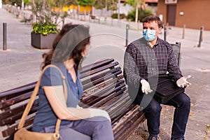 Emotional couple in protective medical masks against coronavirus communicate sittig on bench