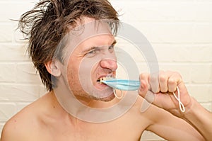 Emotional crazy caucasian man with bristles and shaggy hair, on a white brick background bites and torments medical mask