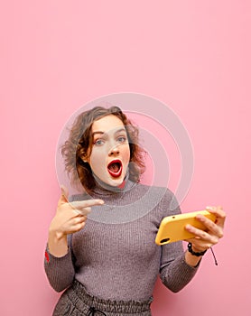 Emotional curly redhead girl in gray stylish casual clothes shows finger on smartphone in hand and looks into camera with