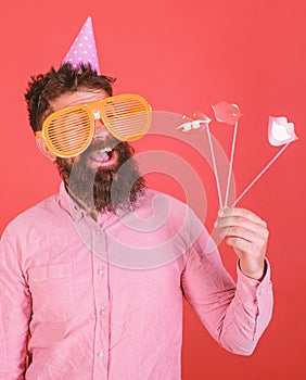 Emotional diversity concept. Man with beard on cheerful face holds smiling lips on sticks, red background. Guy in party