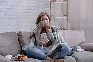 Emotional eating concept. Upset young woman with sweet pastry looking at smartphone, waiting for important message