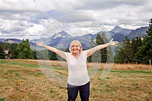 Emotional elderly woman stands among mountains with outstretched arms. Active aging, inner peace