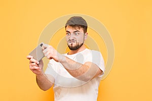 Emotional evil man with a beard playing mobile games on a smartphone, focused on looking at the screen on a yellow background.