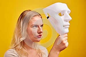 Emotional female person with white mask on yellow background. Internet fraud concept, anonymous, incognito, bipolar personality di