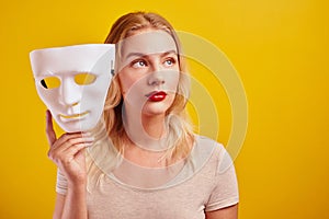 Emotional female person with white mask on yellow background. Internet fraud concept, anonymous, incognito, bipolar personality di