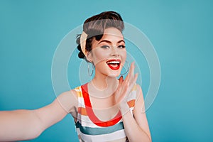Emotional girl in striped dress looking at camera. Studio shot of pretty pinup woman taking selfie on blue background