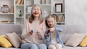 Emotional grandmother and granddaughter playing video games on gamepads, having fun. The little granddaughter wins the