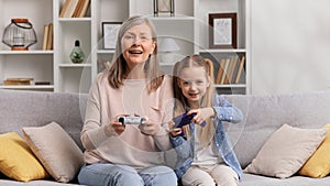 Emotional grandmother in her 60s and young granddaughter playing video games on gamepads, having fun. Senior woman