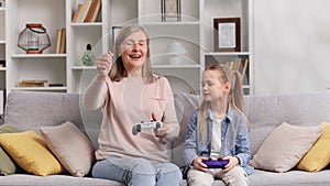 Emotional grandmother in her 60s and young granddaughter playing video games on gamepads, having fun. Senior woman