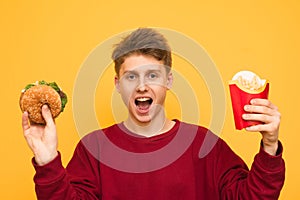 Emotional guy in casual clothes poses on a yellow background with burger and French fries in his hands, looking into the camera.