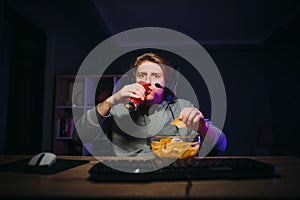Emotional guy gamer with a shocked face playing online games on the computer, drinking soda and eating chips from a plate at home