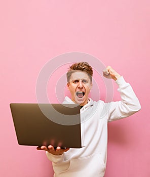 Emotional guy rejoices in victory with a laptop in his hands, looks into the camera and shouts with joy,isolated on pink