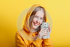 Emotional happy teenage blonde girl in hood win money cash holding dollars in hands isolated on color yellow background. Portrait