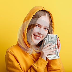 Emotional happy teenage blonde girl in hood win money cash holding dollars in hands isolated on color yellow background. Portrait