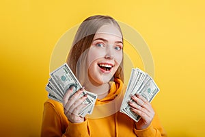 Emotional happy teenage blonde girl win money cash holding dollars in hands isolated on color yellow background. Portrait young