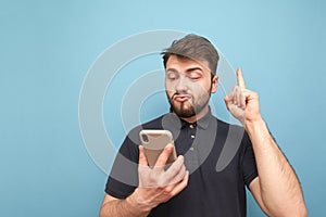 Emotional man with a beard holds a smartphone in his hands, showing his finger up in a blank space. Surprised adult in a dark T-