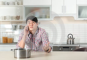 Emotional man calling plumber near table with saucepan under leaking water from ceiling