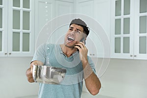 Emotional man calling roof repair service while collecting leaking water from ceiling in kitchen