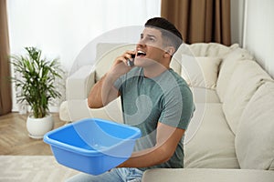 Emotional man calling roof repair service while collecting leaking water from ceiling in living room