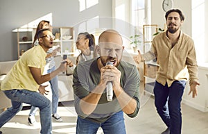 Emotional man holding microphone and enthusiastically singing while having fun with friends at home.