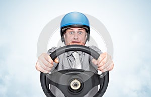 Emotional man in office clothes and motorcycle helmet holding car steering wheel on light blue background, front view.