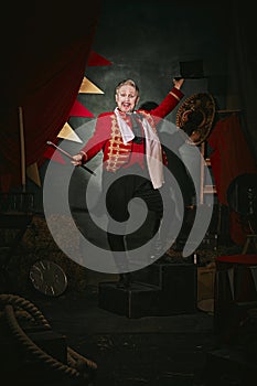 Emotional man with pale face makeup, in red stage costume and hat making performance over dark retro circus backstage