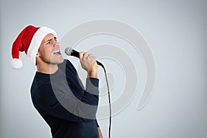 Emotional man in Santa Claus hat singing with microphone on light grey background, space for text. Christmas music