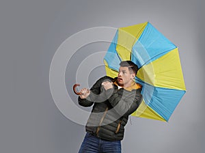 Emotional man with umbrella caught in gust of wind on grey background