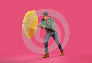 Emotional man with umbrella caught in gust of wind on pink background