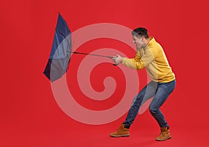 Emotional man with umbrella caught in gust of wind on red background