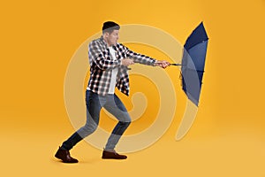 Emotional man with umbrella caught in gust of wind on yellow background