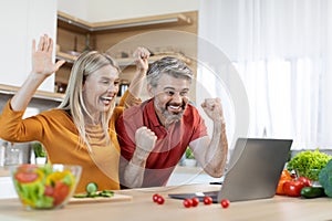 Emotional middle aged couple cooking and using laptop at kitchen