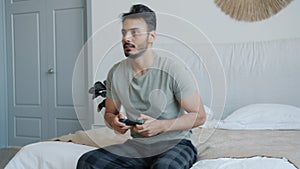 Emotional Middle Eastern man playing video game screaming having fun sitting on bed at home