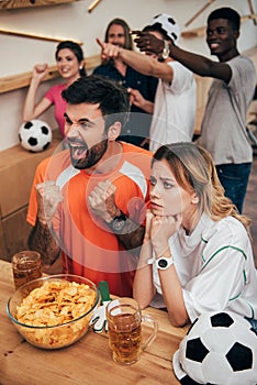 emotional multicultural football fans pointing by fingers and watching soccer match at bar with chips and beer