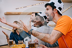 emotional multicultural group of male football fans gesturing by hands and watching soccer match