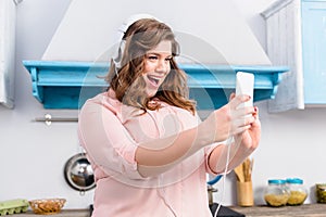 emotional overweight woman in headphones taking selfie on smartphone in kitchen