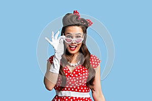 Emotional pin up woman in retro fashion dress touching sunglasses, looking at camera in excitement on blue background