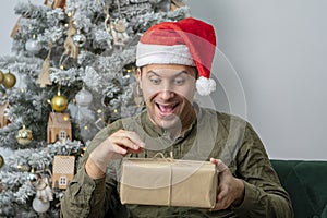 Emotional portrait of a 30-35-year-old man unpacking a gift box on the background of a Christmas tree, a place for your text, copy