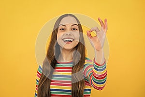 Emotional portrait of caucasian teen girl. Funny teenager making ok gesture, isolated on yellow background. Child