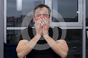 Emotional portrait of an elderly man in a black T-shirt covering his mouth with two hands on a neutral background.Concept: surpris