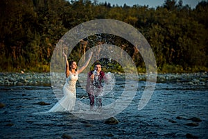 Emotional portrait of the happy fashionable newlyweds splashing water while standing in the river during the sunset.