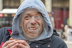 Emotional portrait of a homeless man 45-50 years old in a hood with a mug in his hands on the street, close-up.