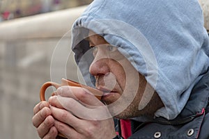 Emotional portrait of a homeless man 45-50 years old in a hood with a mug in his hands on the street, close-up.