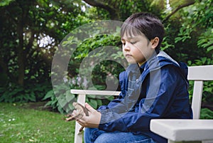 Emotional portrait of Lonely kid sitting alone on bench in the garden, Sad child holding wood stick and looking down deep in