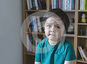 Emotional portrait sad kid looking at camera with unhappy face, Angery little boy sitting alone with blurry bookshelf background,