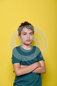 Emotional portrait of a sorrowful, overwhelmed school boy expressing sad emotions, with arms folded on yellow background