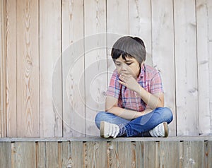 Emotional portrait of Unhappy boy sitting next to wooden wall with sad face, Upset kid sitting alone and looking down with