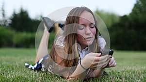 Emotional teenage girl lying on the green grass uses smartphone.